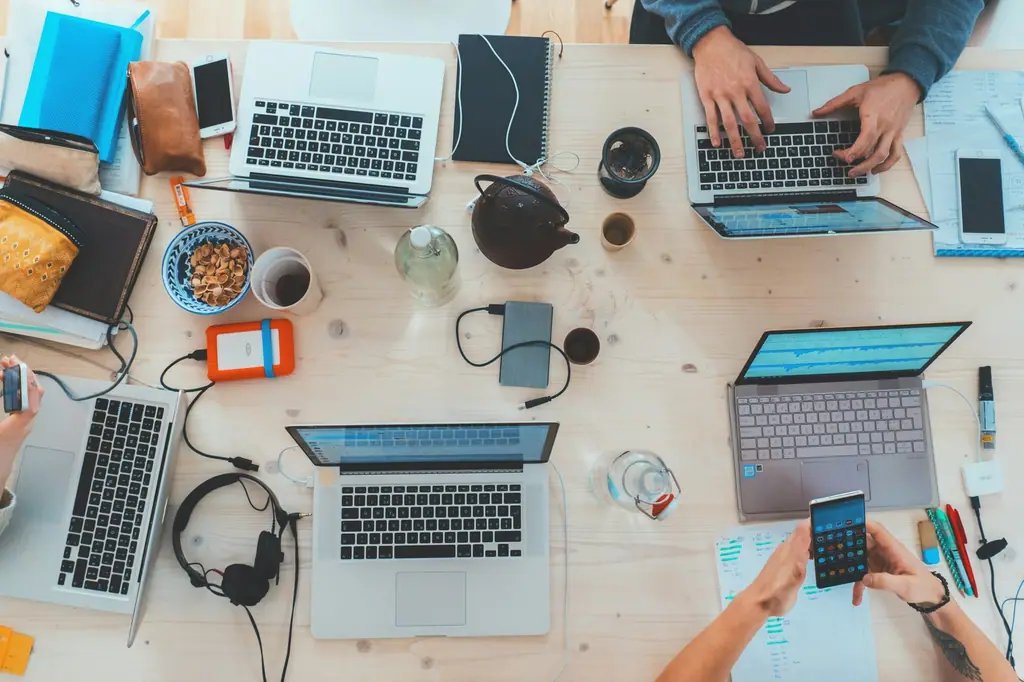 An overhead view of a shared desk with several laptops, phones and notebooks